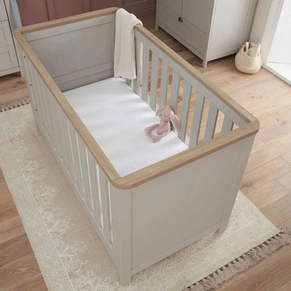 White cot bed with dove grey paneling, oak-toned top rails, and vertical safety slats