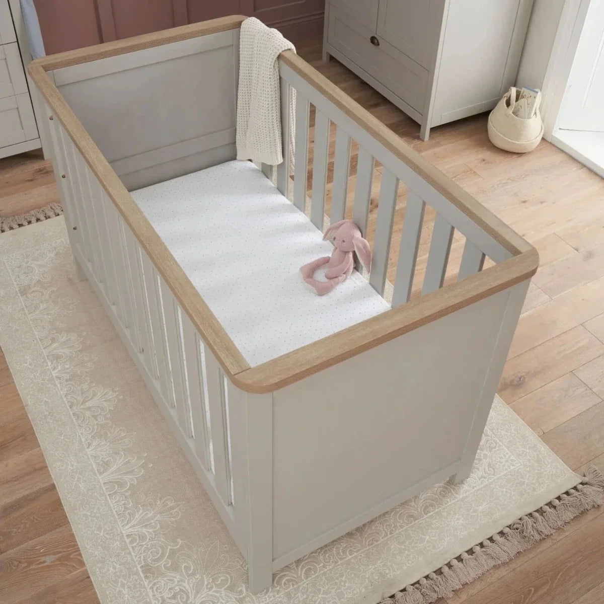 White cot bed with dove grey paneling, oak-toned top rails, and vertical safety slats