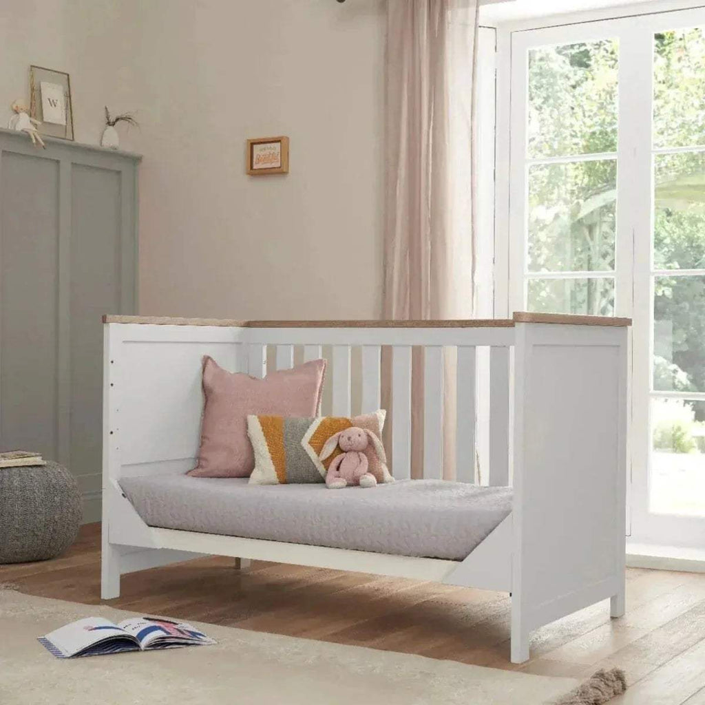 White cot bed with slatted side panels and oak top rail, styled with cushions
