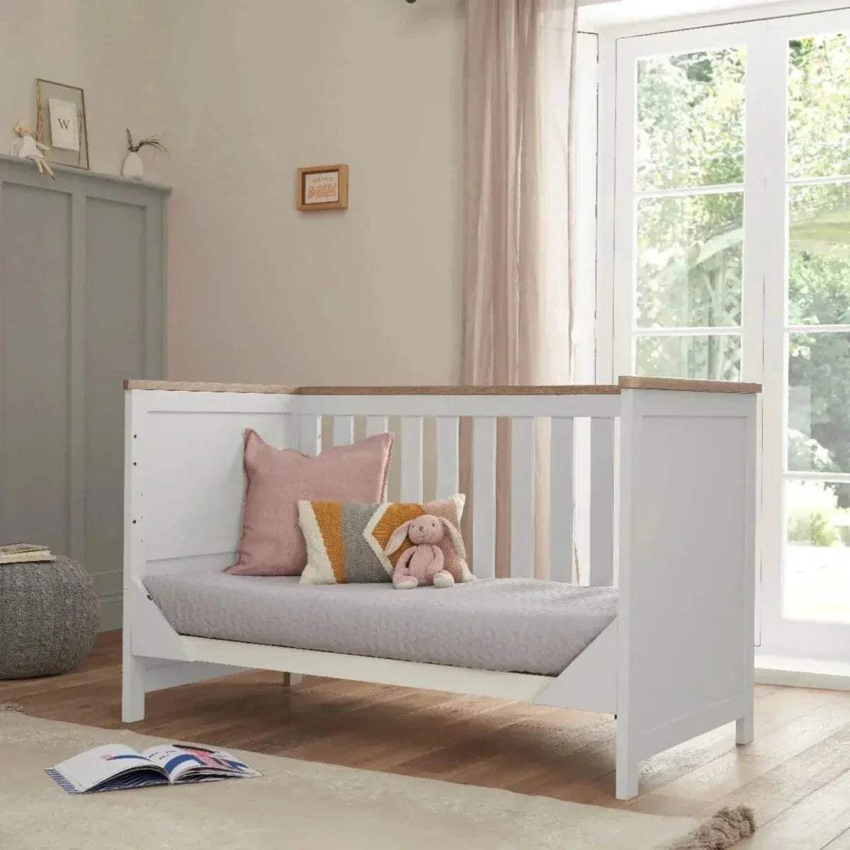 White cot bed with slatted side panels and oak top rail, styled with cushions