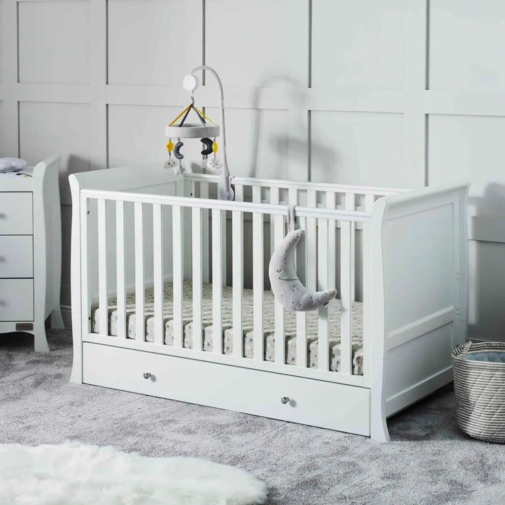 White cot bed with vertical rail, drawer storage front, and mattress inside