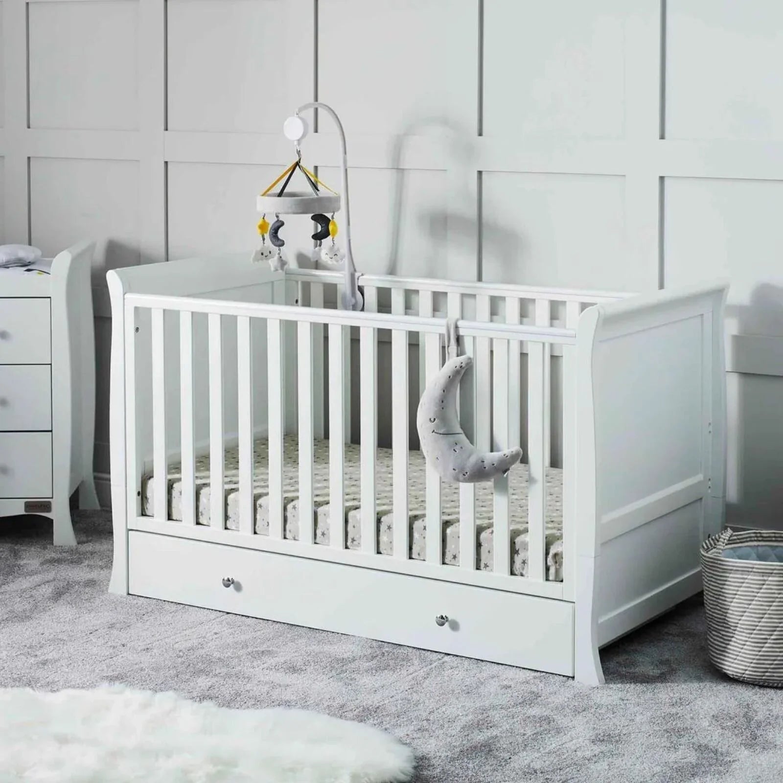 White cot bed with vertical rail, drawer storage front, and mattress inside