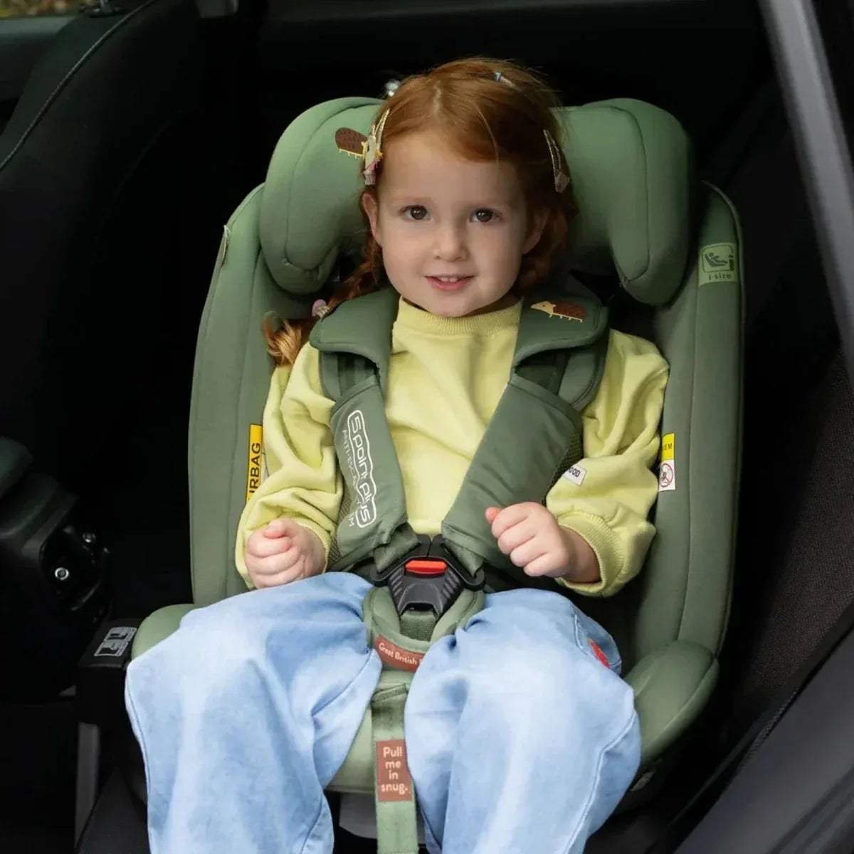 Toddler in green car seat with safety harness and padded headrest.