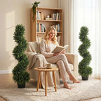 Two deep green artificial cypress trees in black pots near reading chair