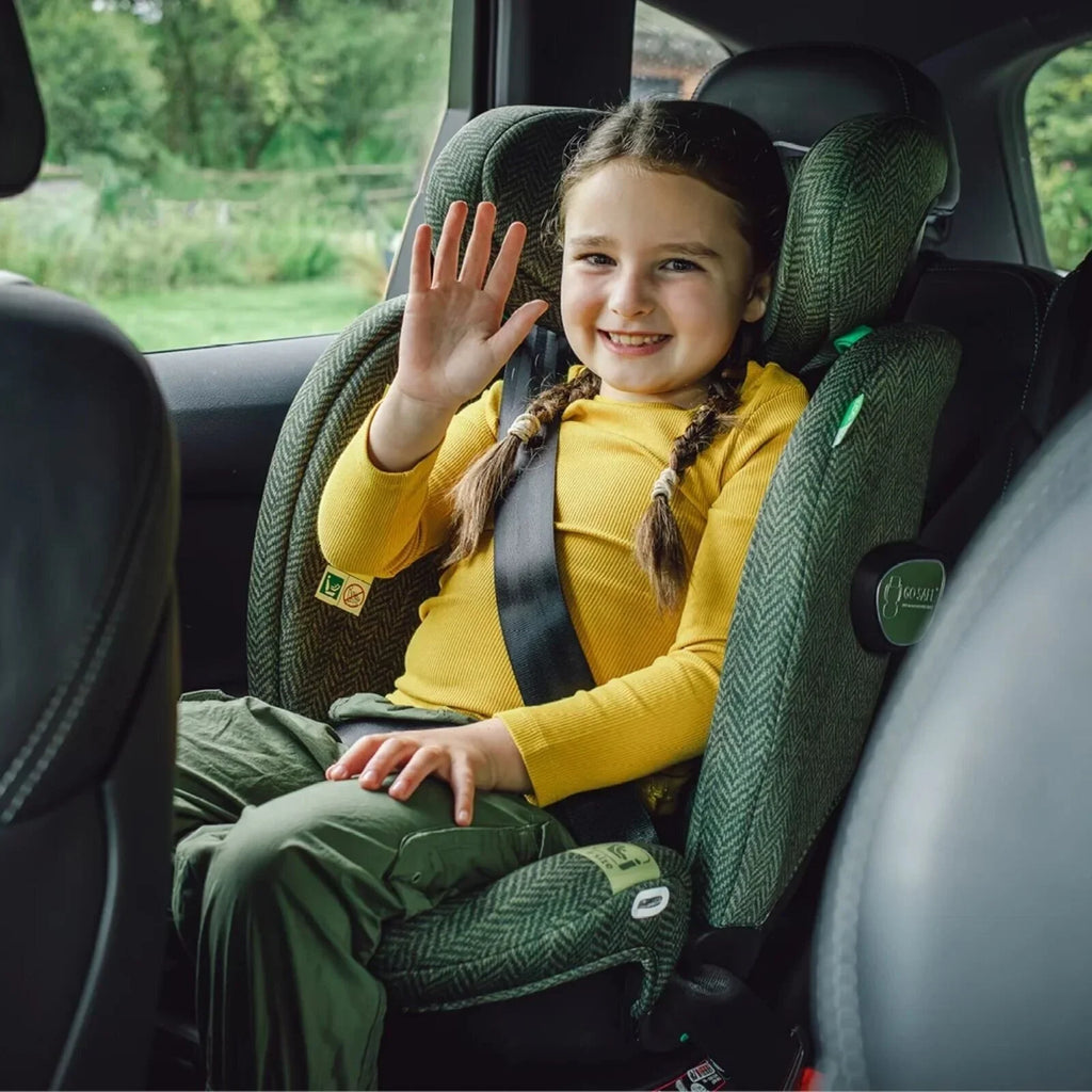 Child in green Cosatto car seat, secured with harness, waving