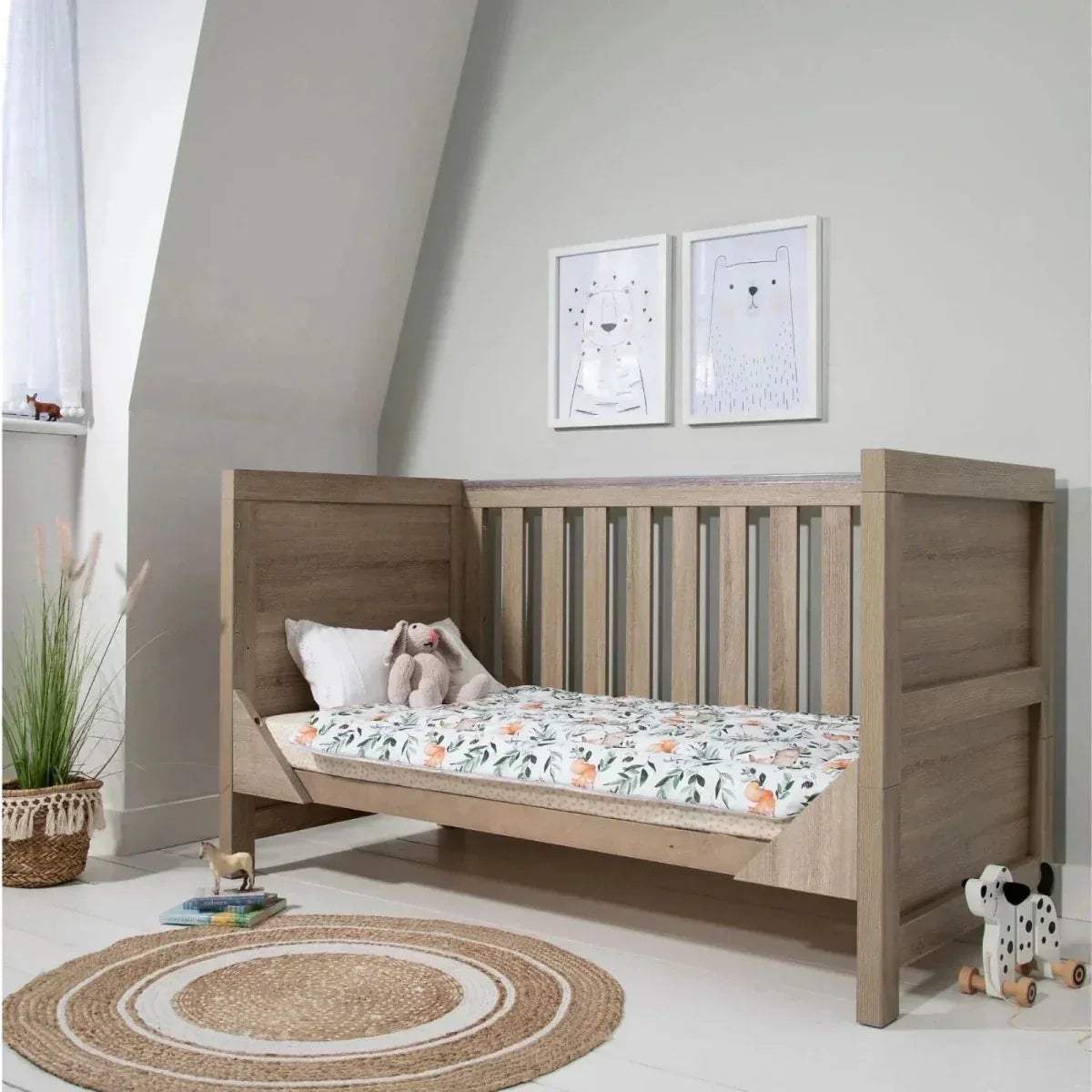 Oak cot bed with white mattress and leaf print fitted cover, slatted sides and end rails