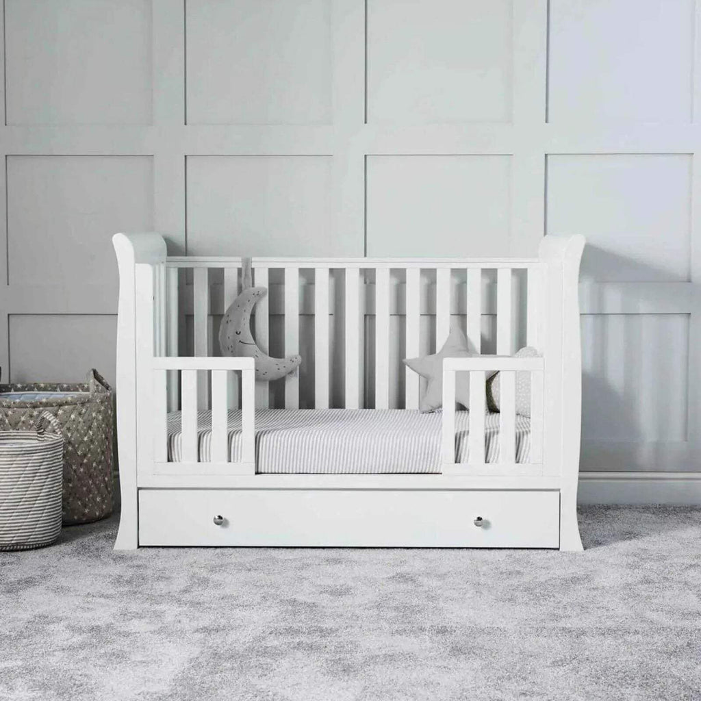White baby crib with striped mattress, decorative pillows, and drawer front