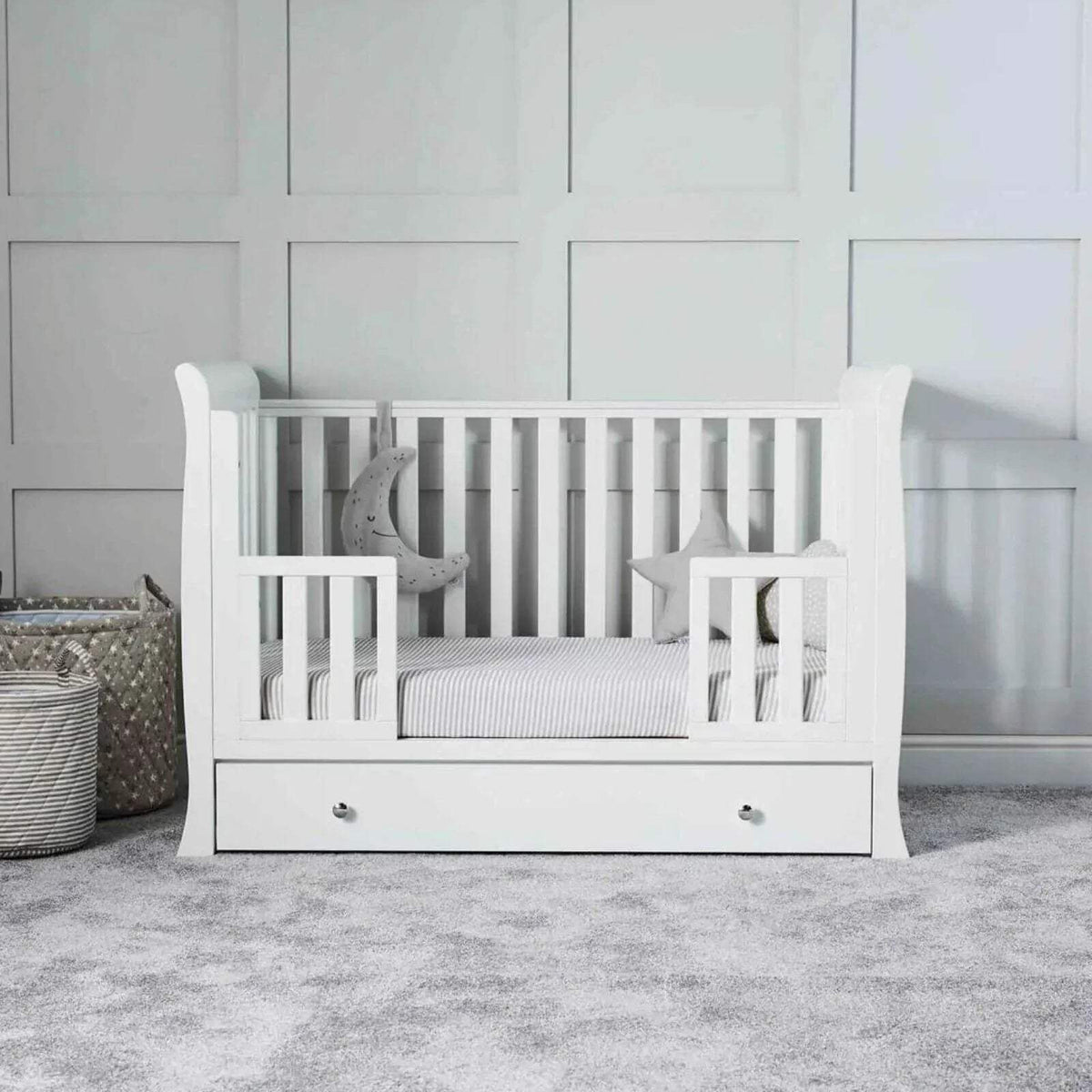 White baby crib with striped mattress, decorative pillows, and drawer front