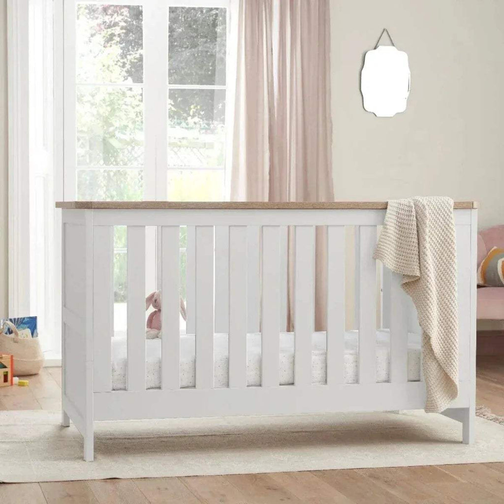 White cot bed with slatted sides and oak top rail, with a beige blanket