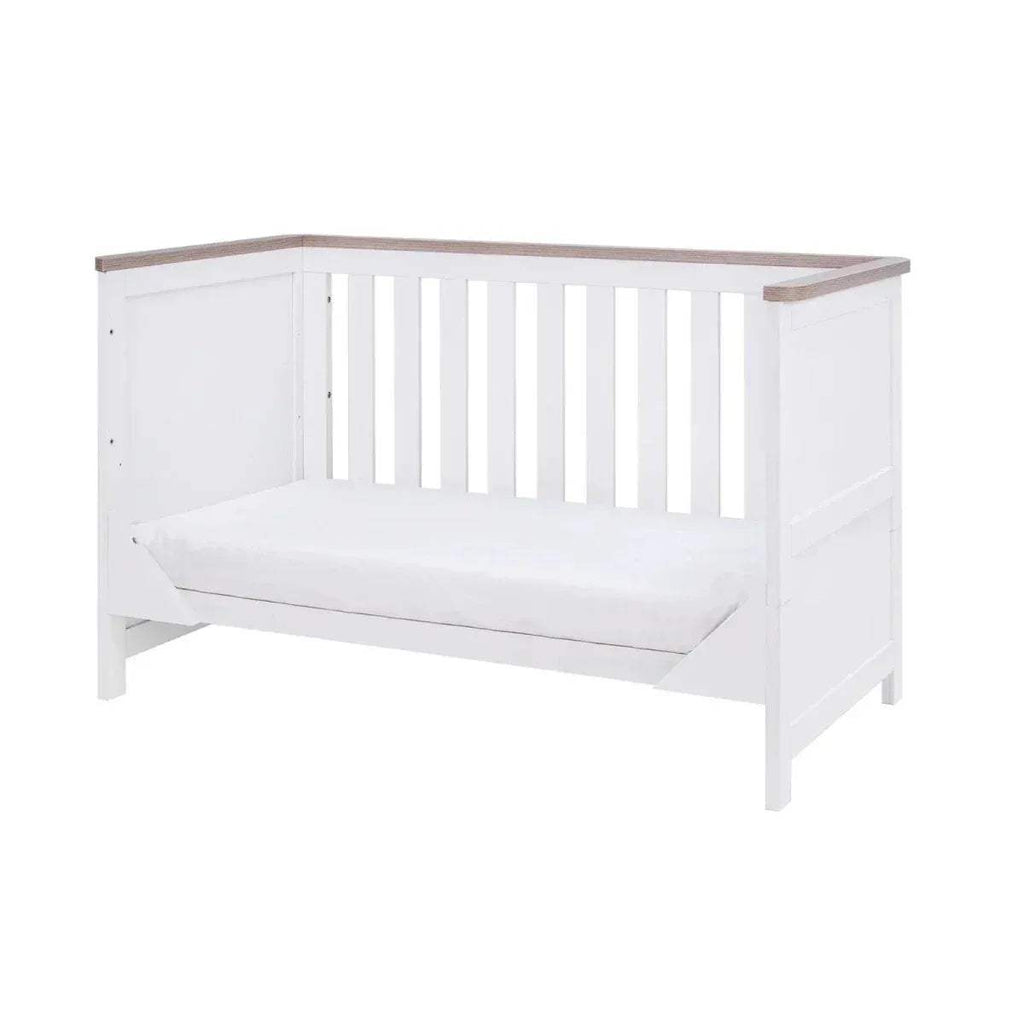 White cot bed with slatted rails and oak top trim, with a white mattress
