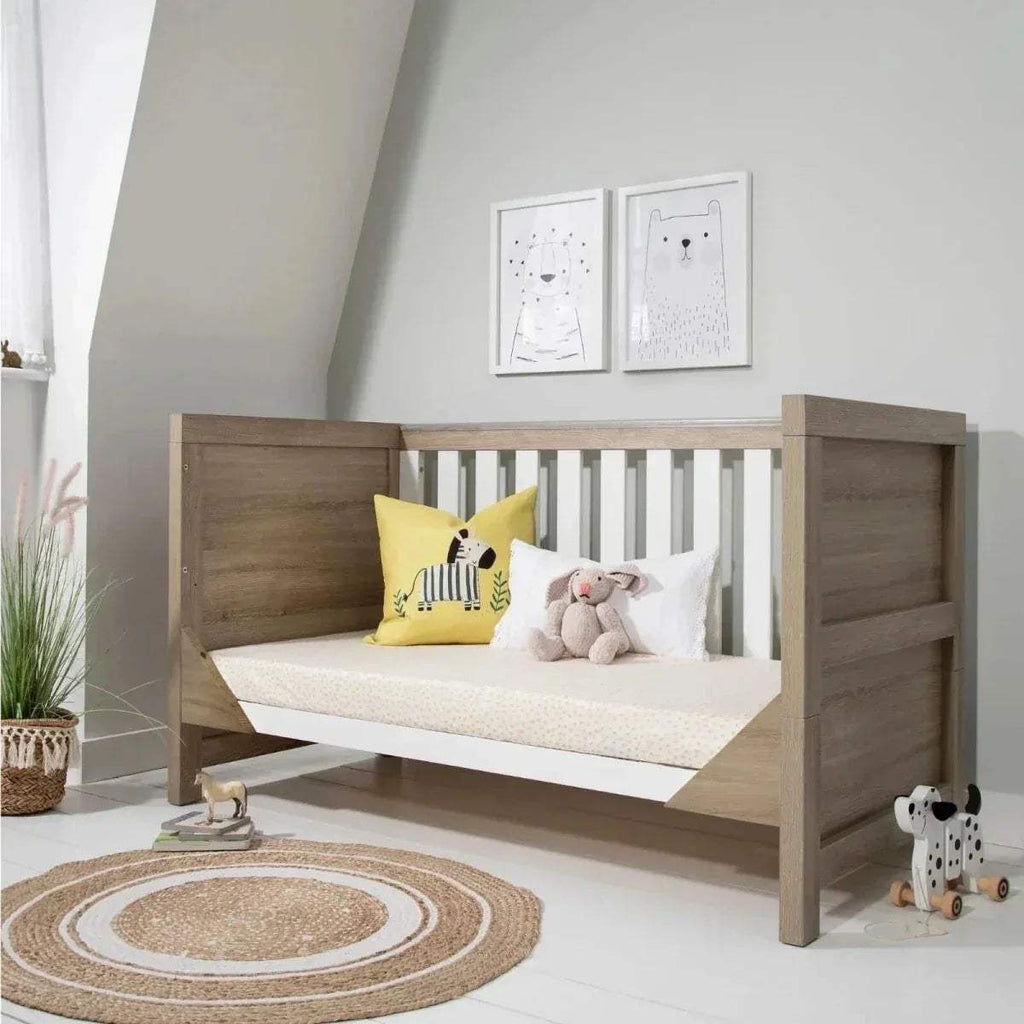 Light oak cot bed with white slats and patterned bedding, with pillows.