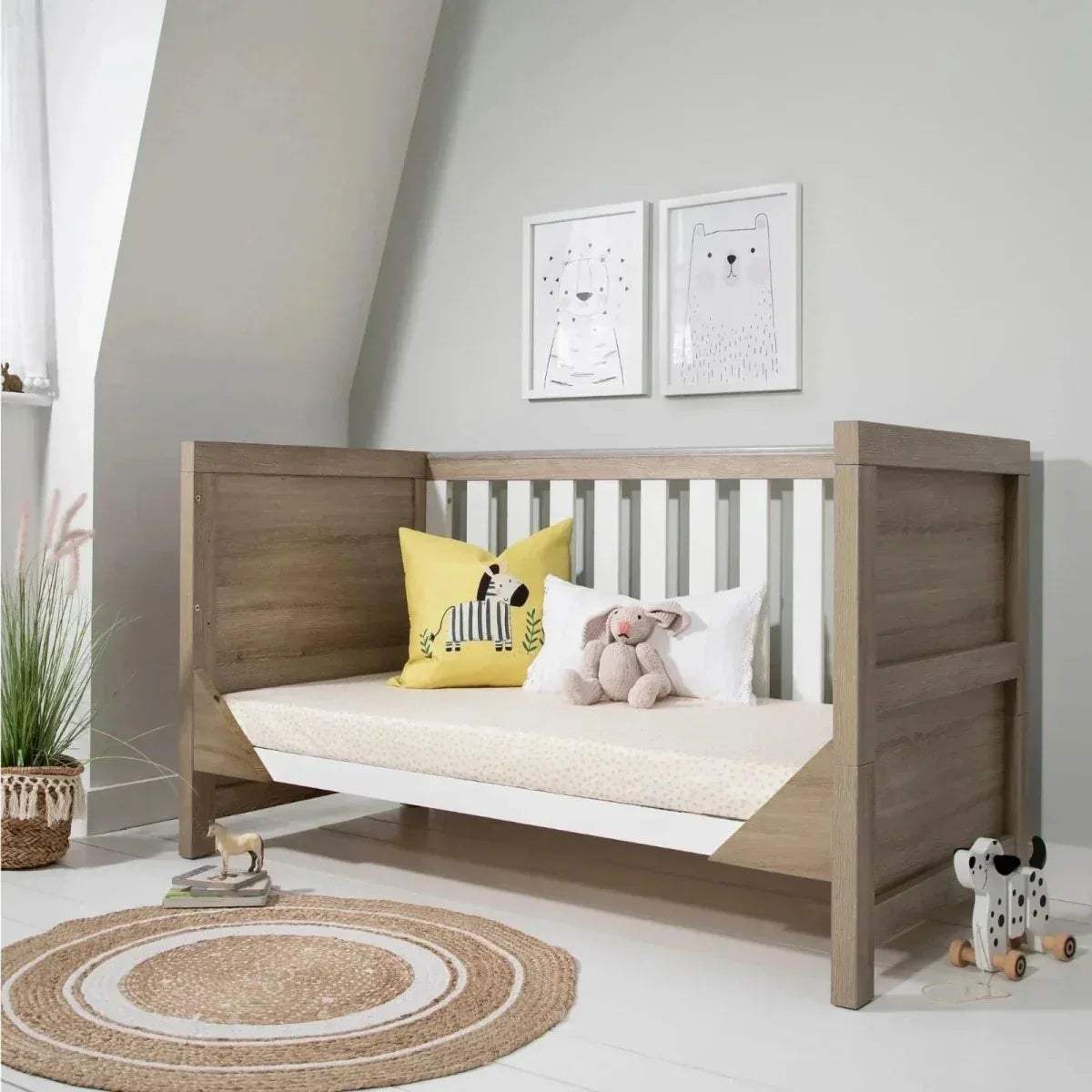 Light oak cot bed with white slats and patterned bedding, with pillows.