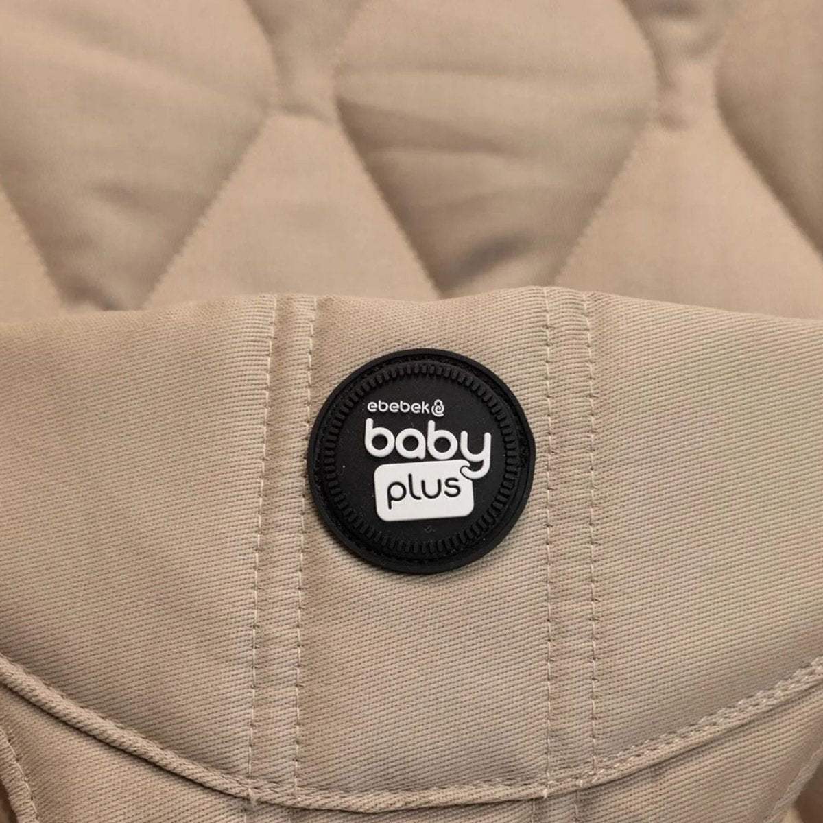 Beige bouncer seat with black "baby plus" logo patch on fabric