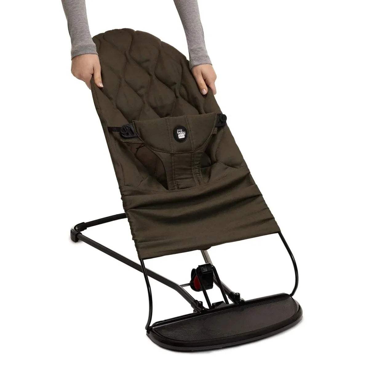 Khaki baby bouncer being held, showing padded seat, harness, and rocking frame.