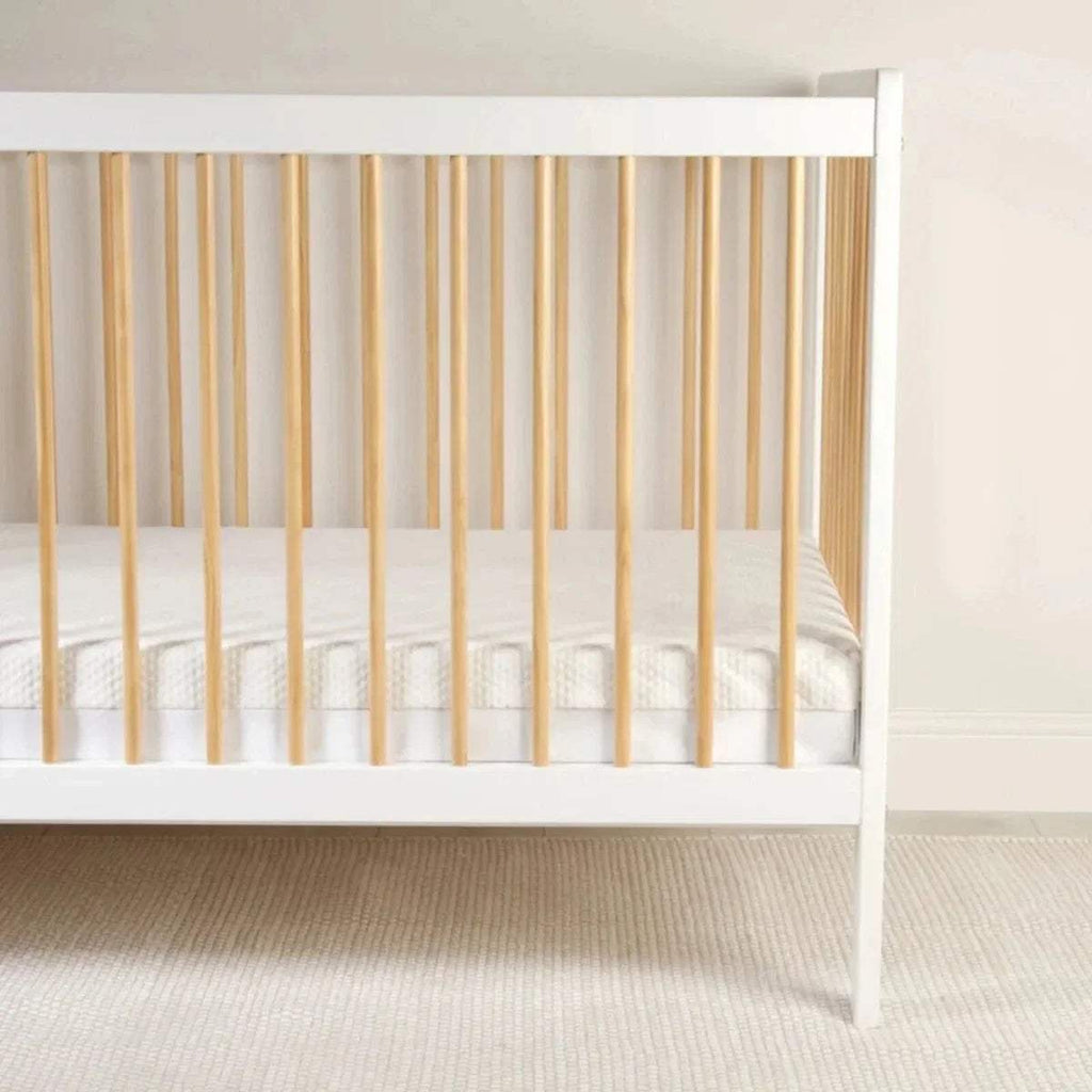 Cot bed mattress with white cover in a white wooden cot, front view.