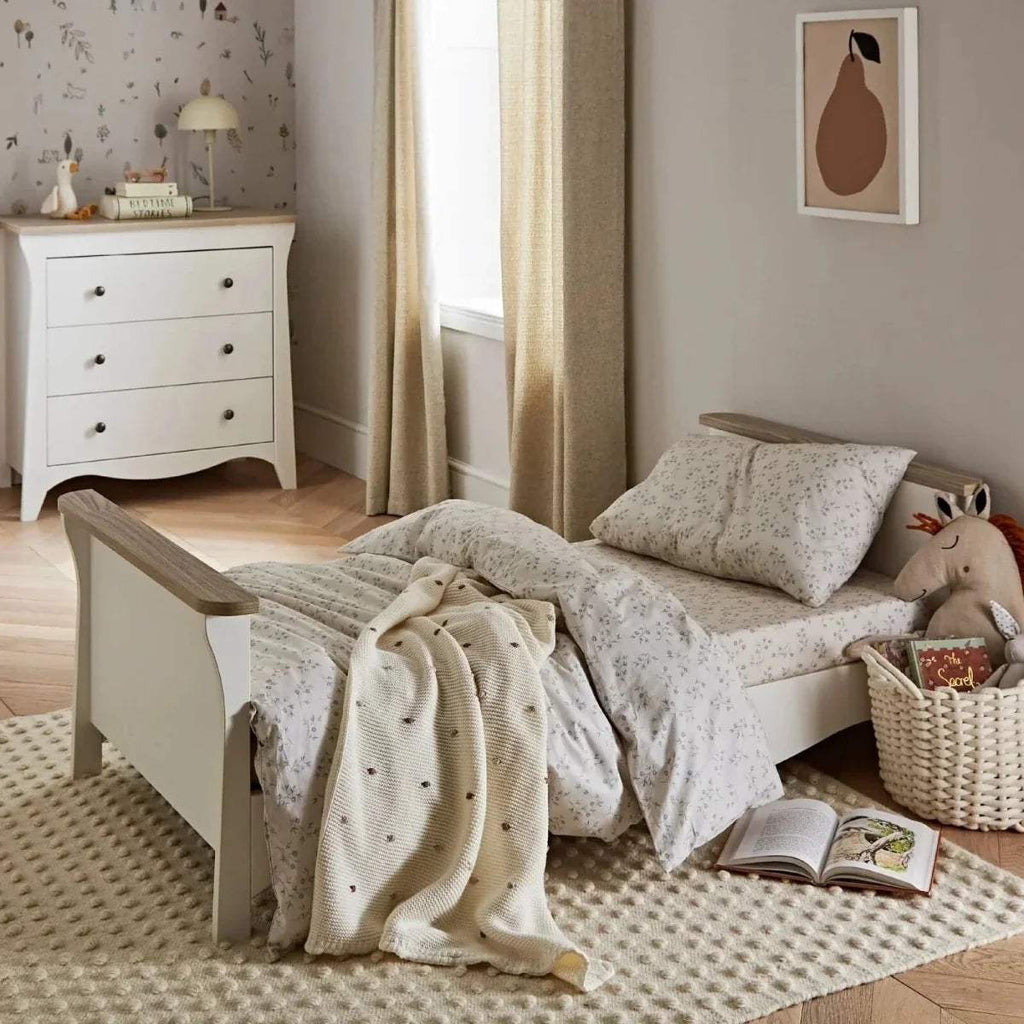 White toddler bed with slatted frame, patterned bedding, pillows, and soft blanket