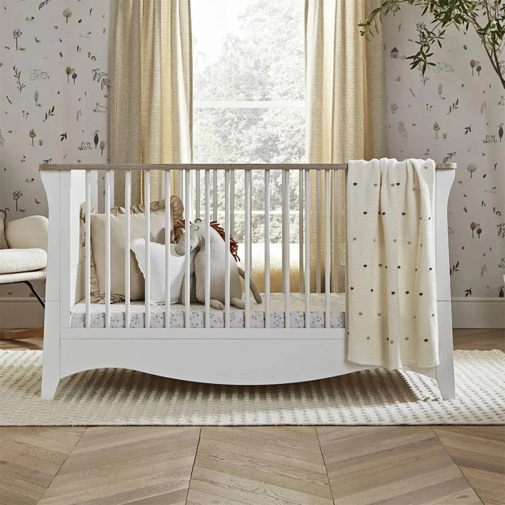 White nursery crib with tall slats and ash-toned feet; cream blanket on mattress.