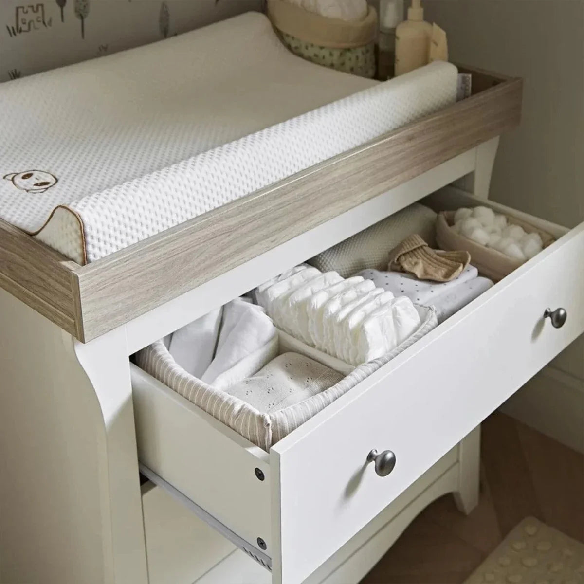 White nursery bed with ash side rail and pull-out drawer for folded bedding.