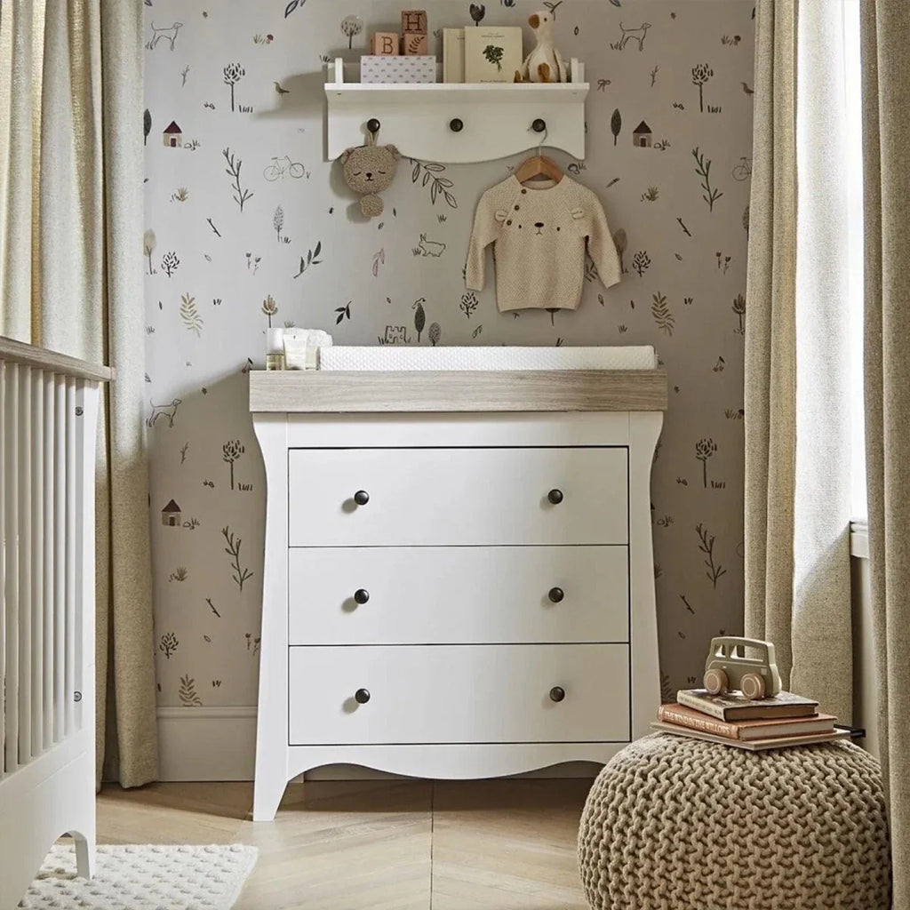 White nursery dresser with three drawers and changing ledge; wall-mounted clothes above.