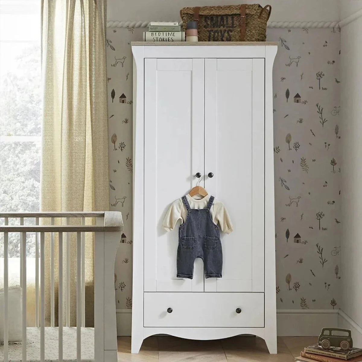 White nursery wardrobe with double doors and lower drawers; hanging baby outfit.