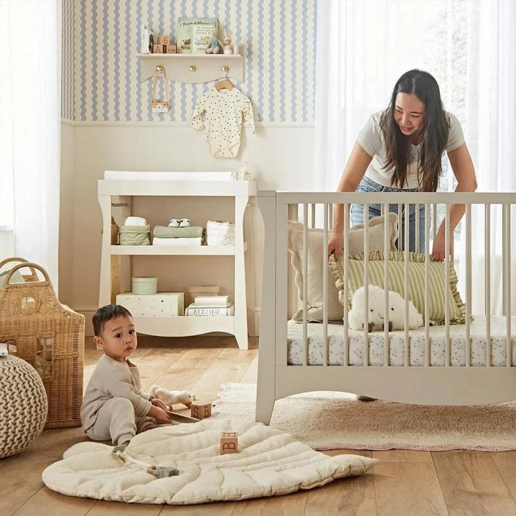 White nursery crib with slatted sides, padded bedding and baby on play mat