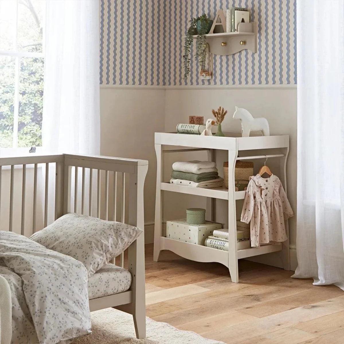 White storage dresser with shelves, toys, and hanging baby outfit next to crib