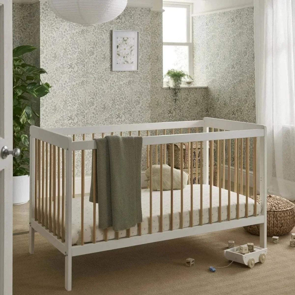 White and natural cot bed with vertical slats, raised sides, and grey fabric blanket.