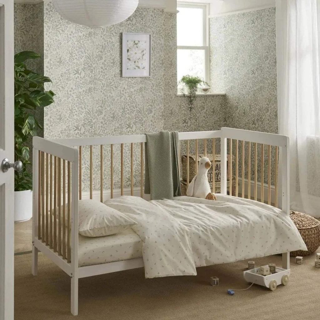 White and natural cot bed with slatted sides, grey cover, and small plush toy on mattress.