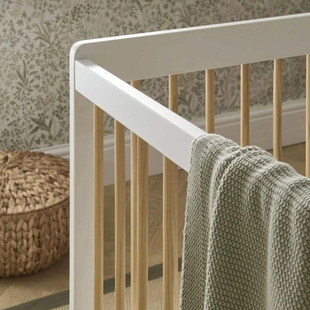 Close-up of white cot bed rail and natural slats, with textured grey blanket draped.