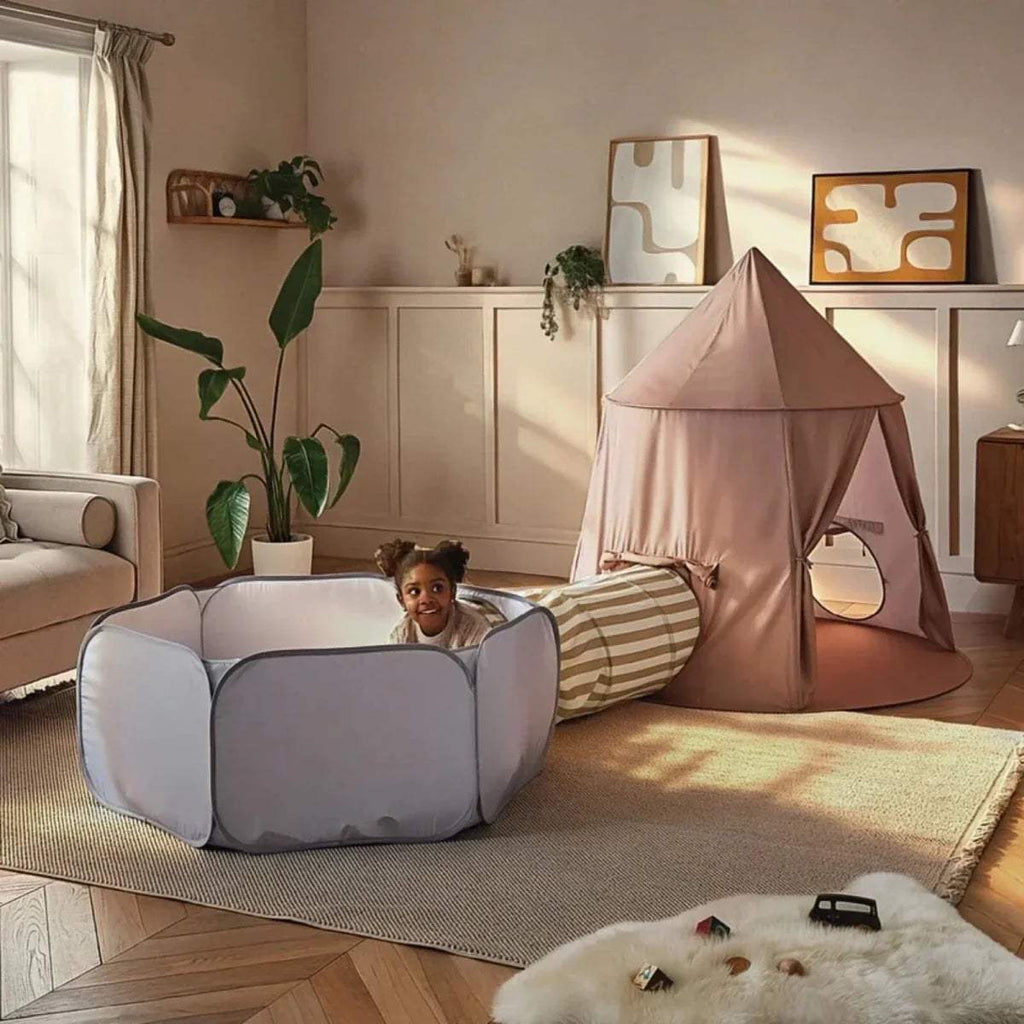 Child playing beside a dusty pink play tent and light gray padded ball pit with striped tunnel