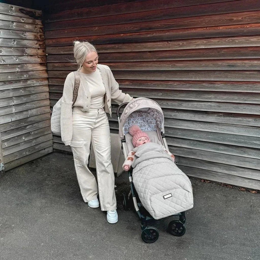 Beige stroller with padded footmuff and baby, outdoors near wooden wall