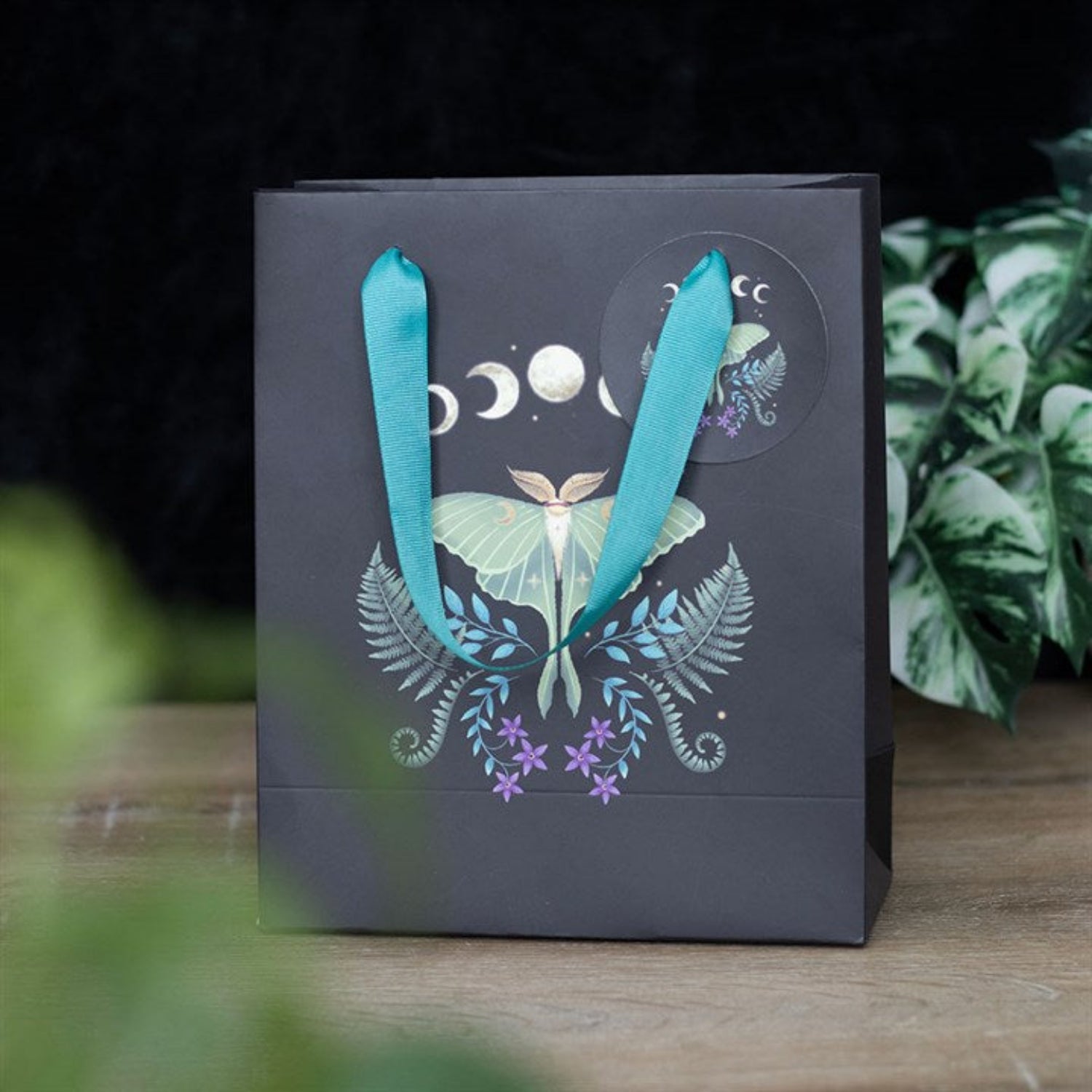 Gift bag with decorative design and blue ribbons on a wooden surface with plants in the background