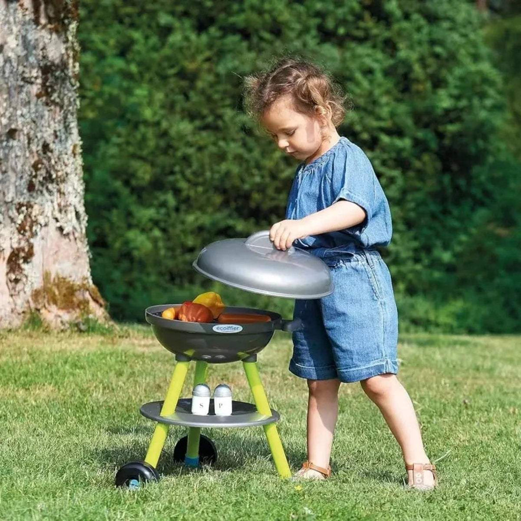 Kids toy BBQ grill with dark lid and lime stand; child places food on the grill