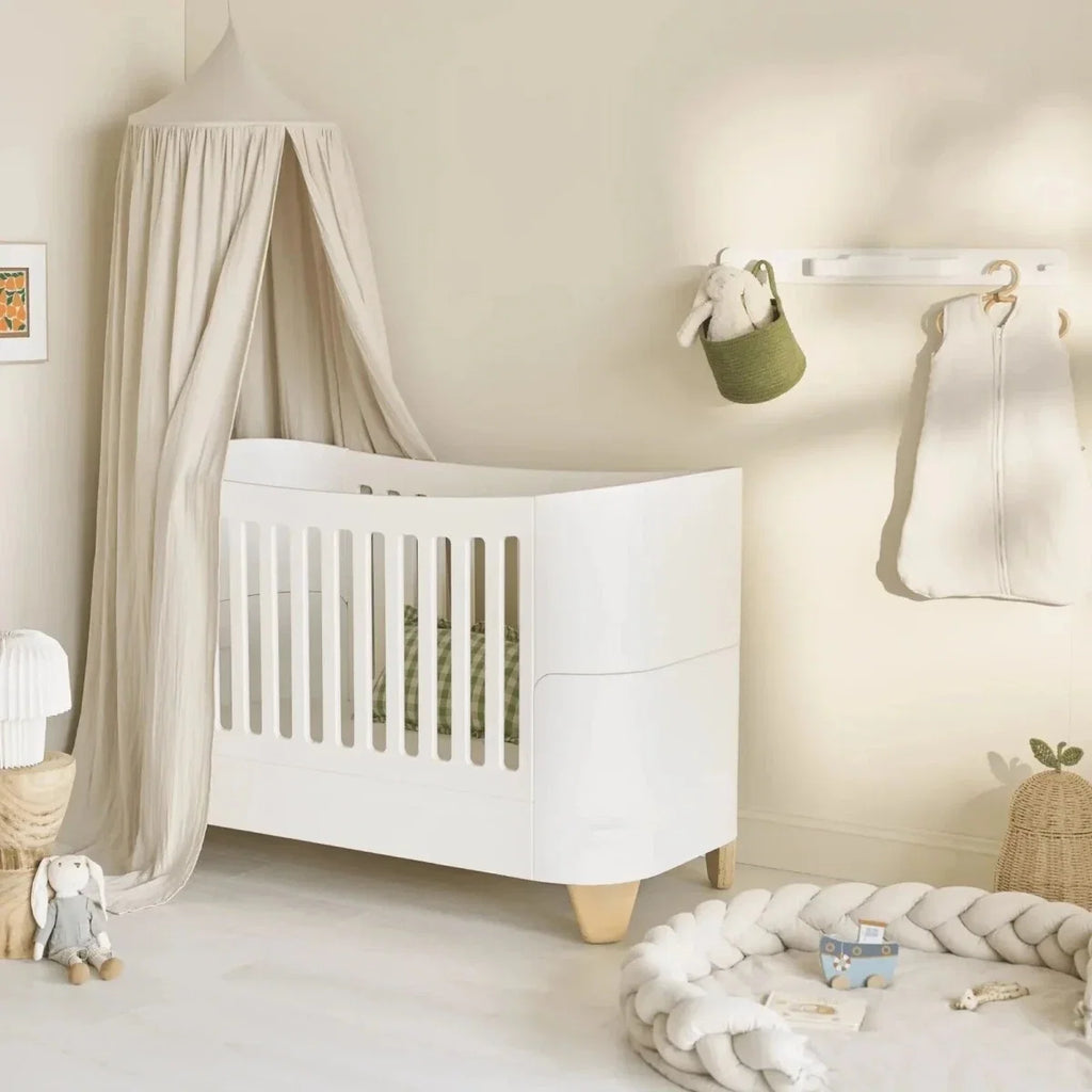 White bedside crib with slatted panels, curved sides, and natural wood legs