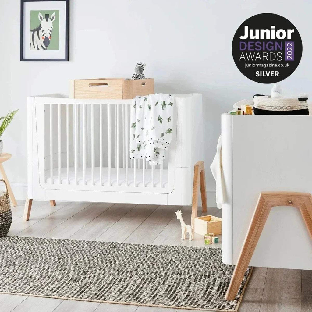 Convertible cot bed with white frame, vertical slats, light wood legs, and safety rail ends