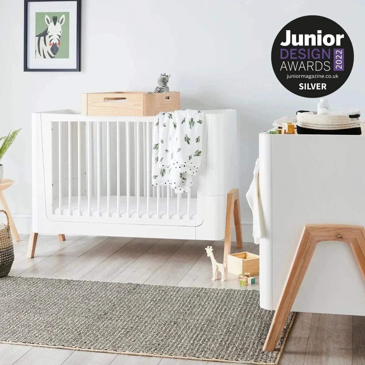 Convertible cot bed with white frame, vertical slats, light wood legs, and safety rail ends
