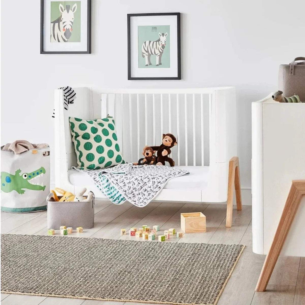 Convertible cot bed in white with vertical rails and light wood legs, shown with fitted bedding