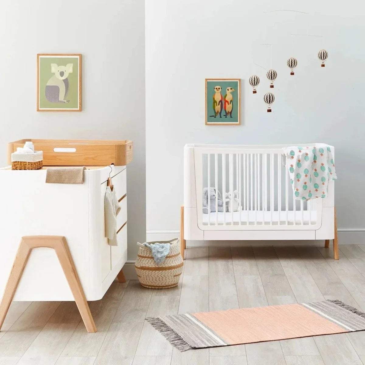 Convertible cot bed with white frame and light wood legs, shown beside changing dresser base