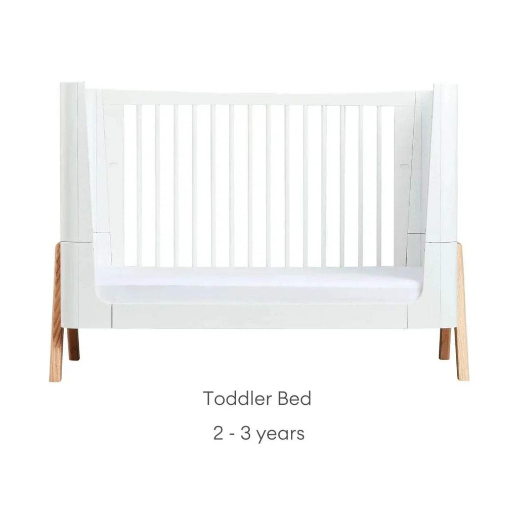 Convertible cot bed in toddler mode with white rail and natural wood legs
