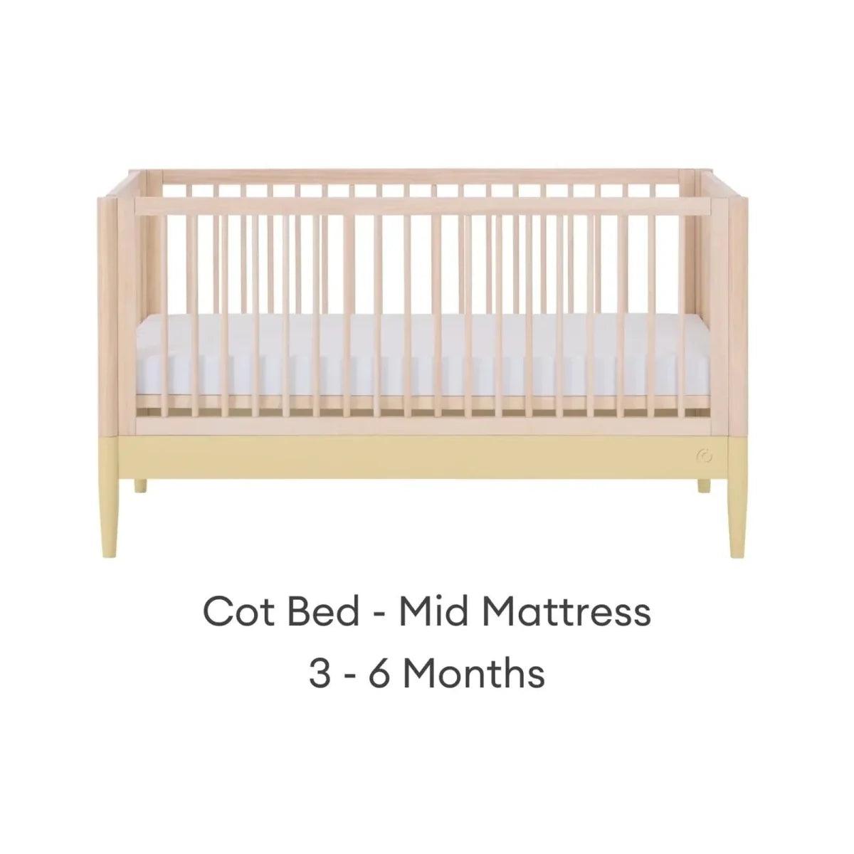 Cot bed with mid mattress level, light wood frame and white mattress panel