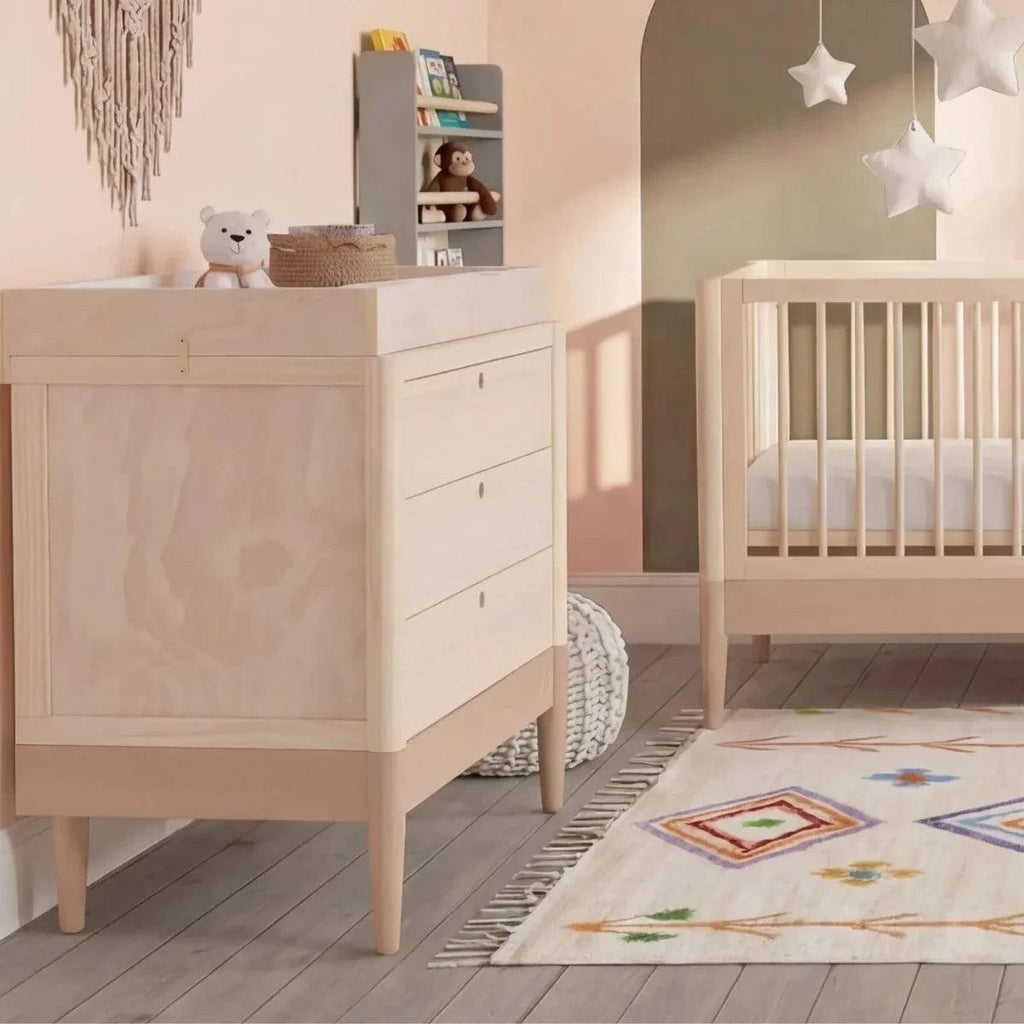 Light wood baby cot bed beside white storage dresser, rug and decor in nursery
