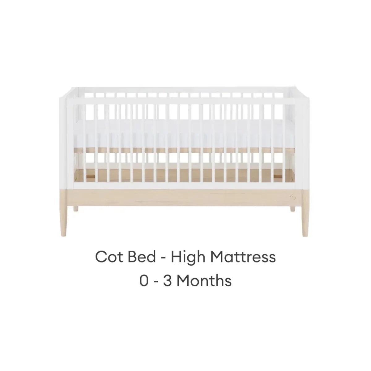 White cot bed with high mattress position, natural wood panel, and slatted rails.