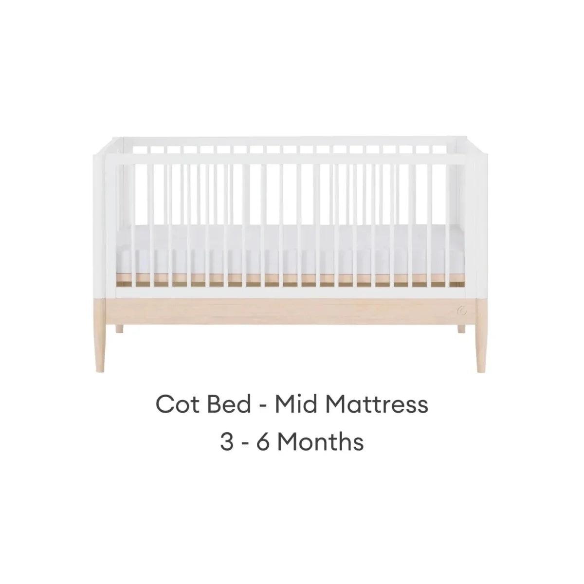 White cot bed with mid mattress position, natural wood panel, and vertical slat rails.