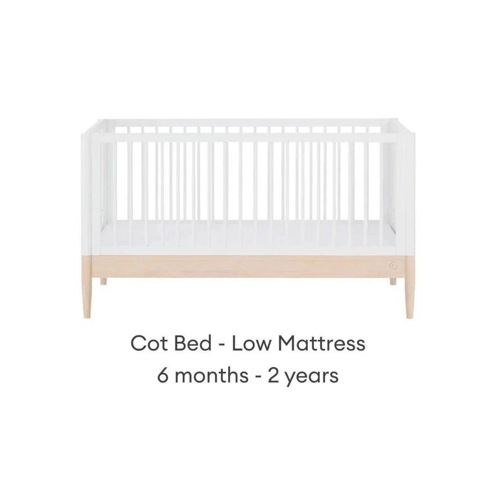 White cot bed with low mattress position, natural wood panel, and side slat rails.