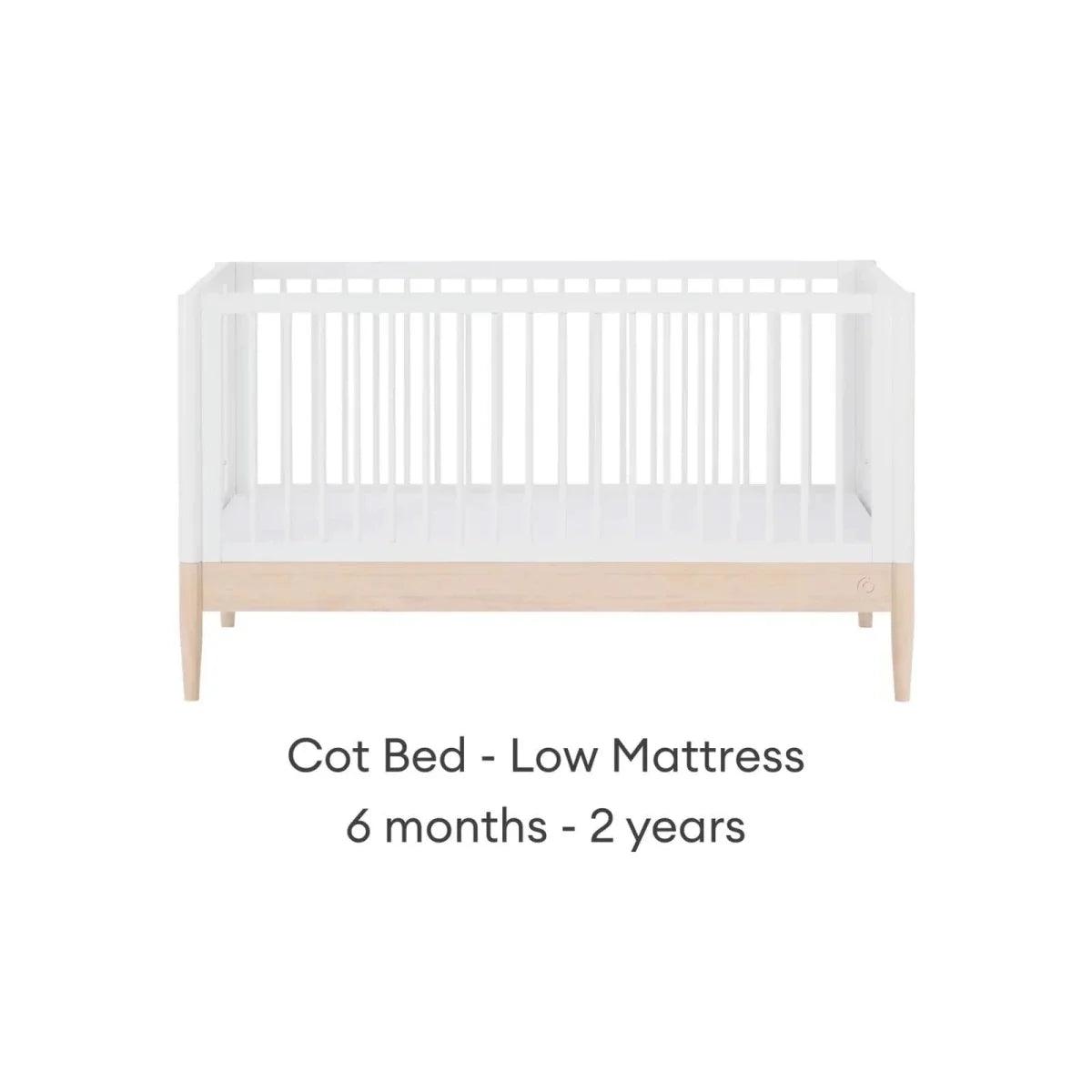 White cot bed with low mattress position, natural wood panel, and side slat rails.