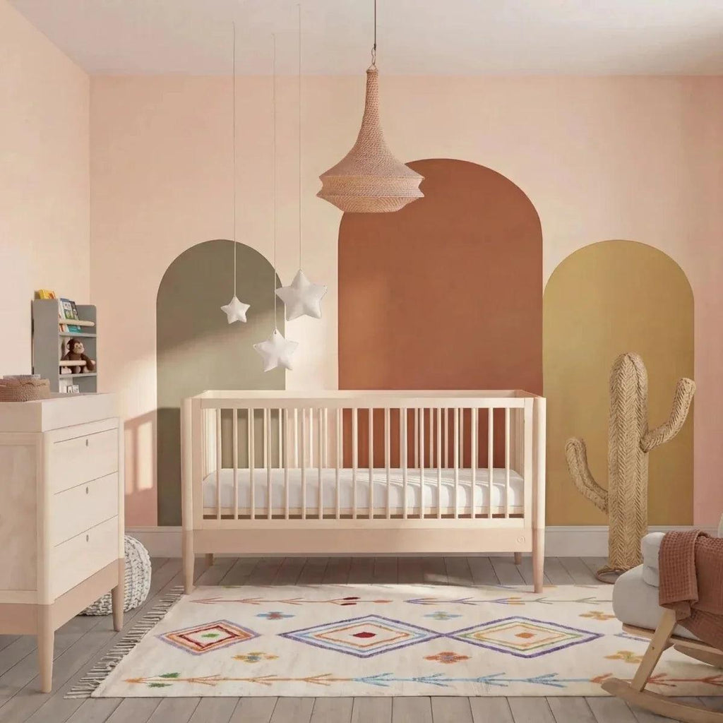Nursery cot bed with powder blush frame and natural ash slatted sides, raised mattress.