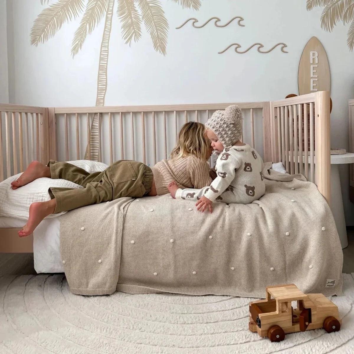 Powder blush and natural ash cot bed with slatted sides, toddler playing on blanket.