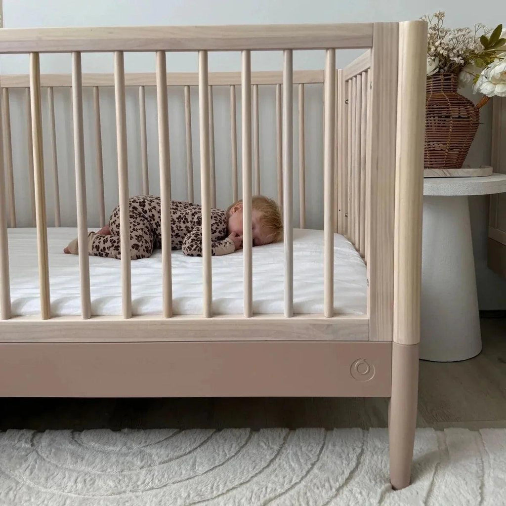 Natural ash cot bed with powder blush base panel and vertical slats, baby lying inside.