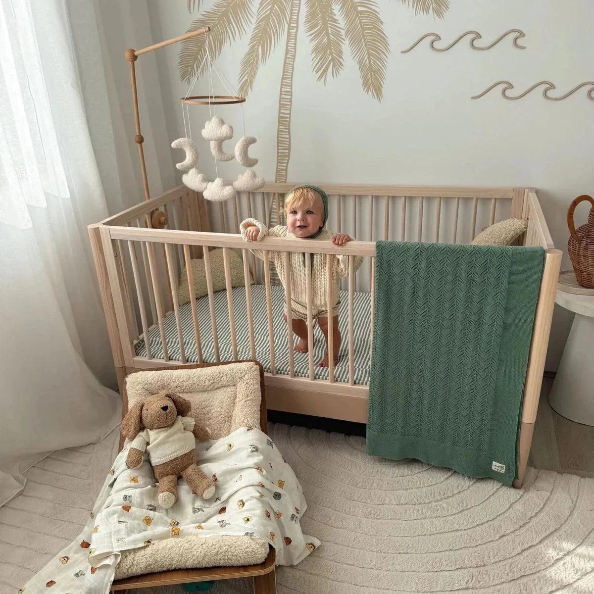 Powder blush cot bed with natural ash rails and slatted sides, baby standing in bed.