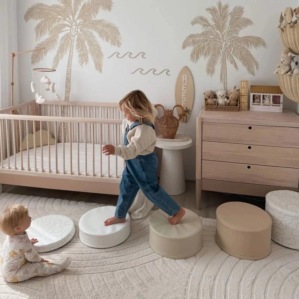Natural ash cot bed with powder blush panels and slats, child standing beside the mattress.
